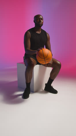 Vertical-Video-Studio-Shot-Of-Seated-Male-Basketball-Player-Bouncing-Ball-Against-Pink-And-Blue-Lit-Background